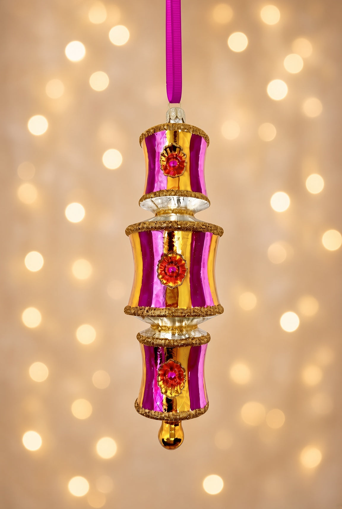 Three-tiered glass ornament in glossy fuchsia and gold vertical stripes with gold glitter trim and round ruby reflector medallions, photographed against a blurred backdrop of warm golden bokeh lights.