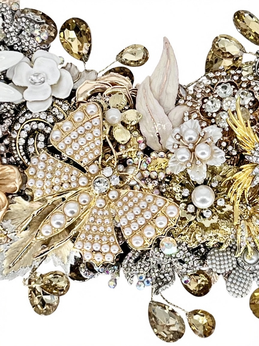 A close-up detail of a jeweled wreath showcasing a golden butterfly in pearl-studded accents, white enamel florals, gold filigree leaves, champagne and aurora borealis accent