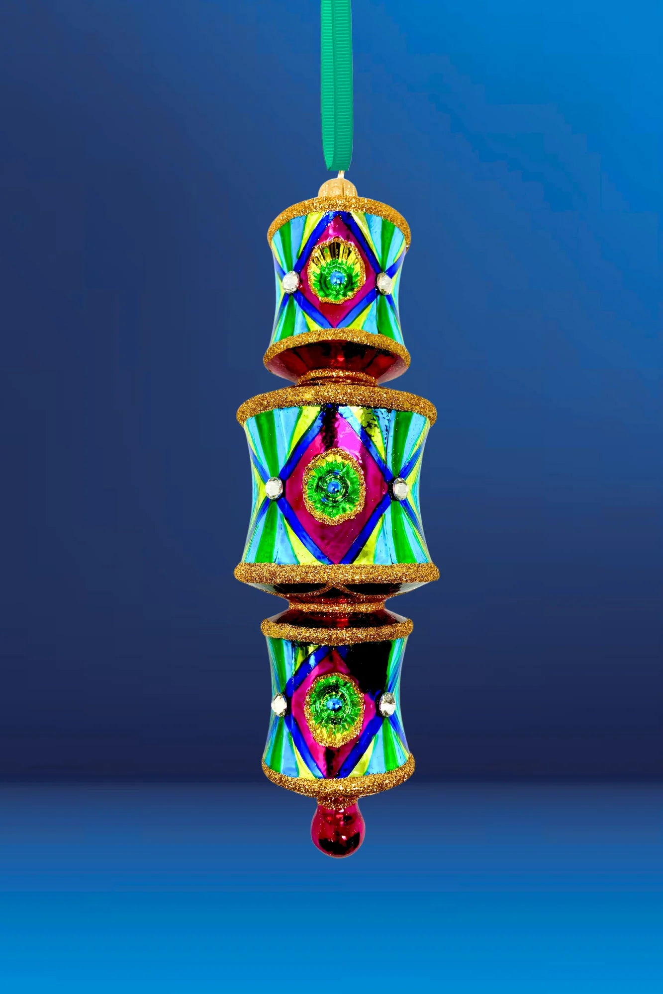 On a blue background a three-tiered barrel-shaped glass ornament with red and gold glitter banding at the top and bottom of each tier, fuchsia front panels centered on blue reflectors casting a green glow onto yellow, faceted crystal accents, alternating yellow-green and blue striped side panels within a dark blue border, red connecting rings between tiers, and a fuchsia teardrop drop.