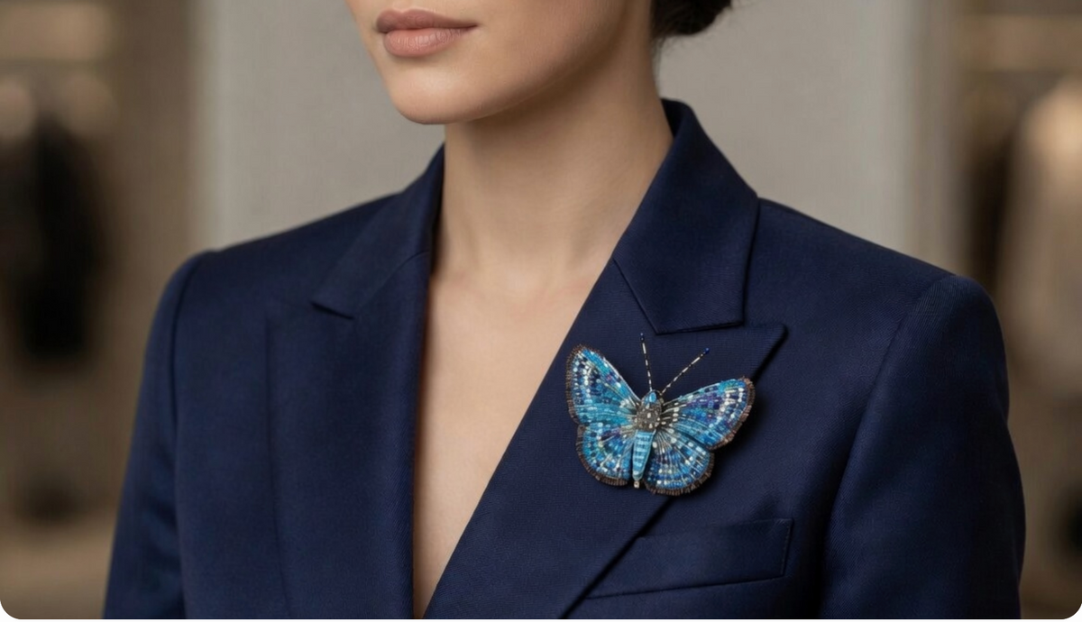 On a womans blue blazer is a Hand-embroidered butterfly brooch pin with wings spread in a mosaic of turquoise, cobalt, and navy bugle beads flecked with white, gold, and magenta, edged in a bronze fringed border, with a ridged turquoise body and gold and black banded antennae tipped in blue.