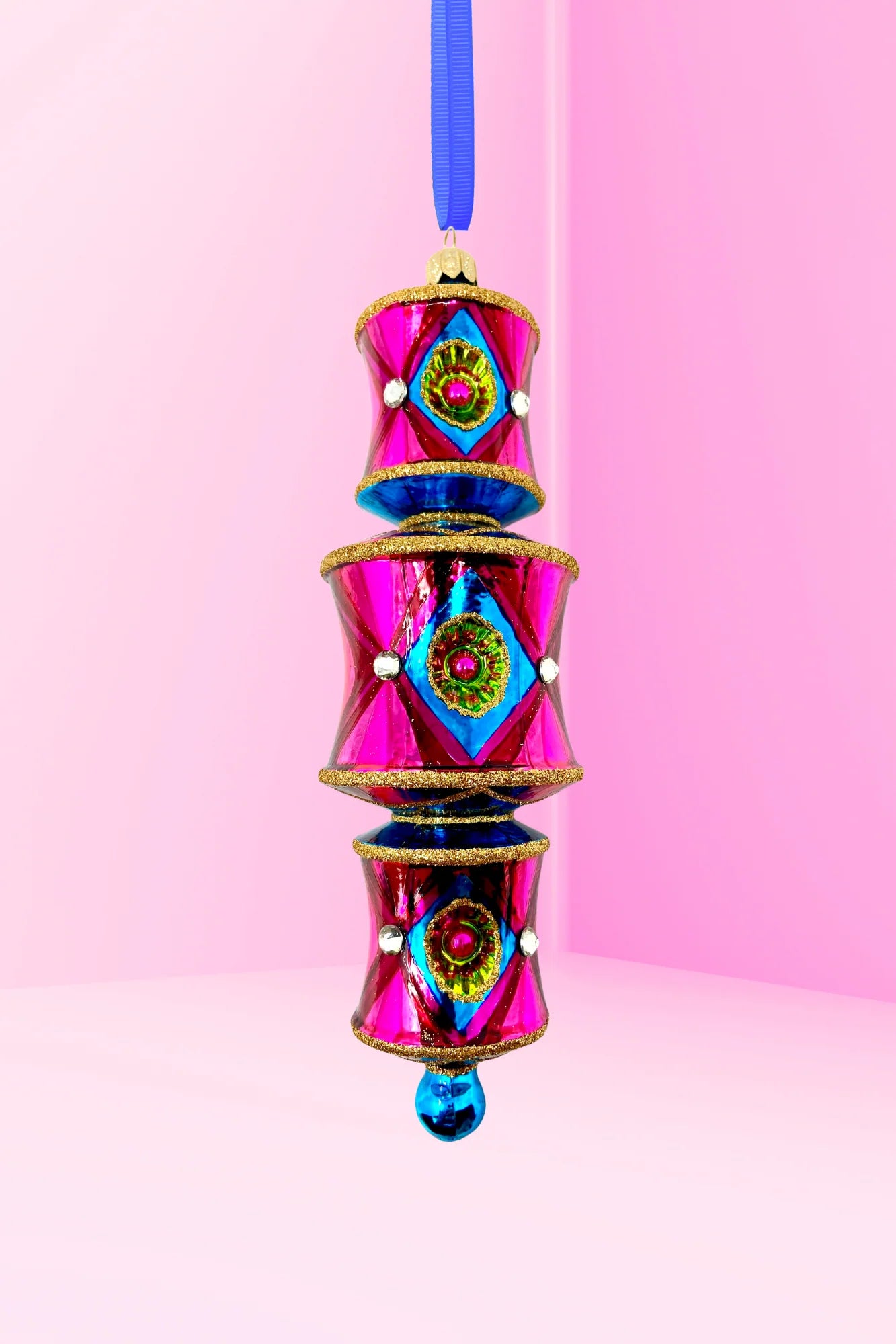On a pink background a three-tiered barrel-shaped glass ornament in hot pink deepening to red, with blue diamond reflectors catching flashes of green and pink, faceted crystal accents, gold glitter trim, and a blue glass drop, hung from a blue ribbon.