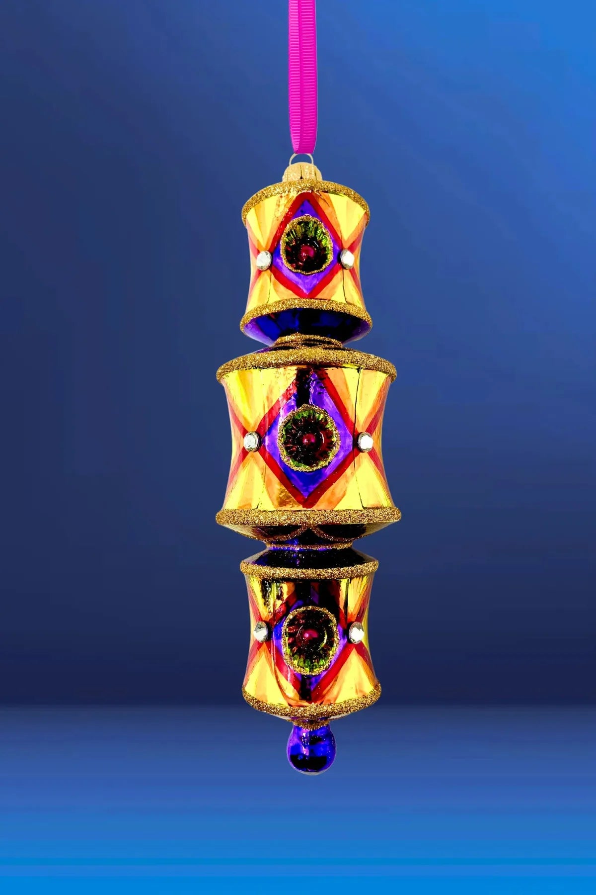 On a blue background is a three-tiered barrel-shaped glass ornament in gold with a red diamond lattice pattern, reflectors banded in violet and green around a maroon center, faceted crystal accents, deep blue-purple connecting sections between tiers, gold glitter trim, and a blue glass drop, hung from a magenta ribbon.