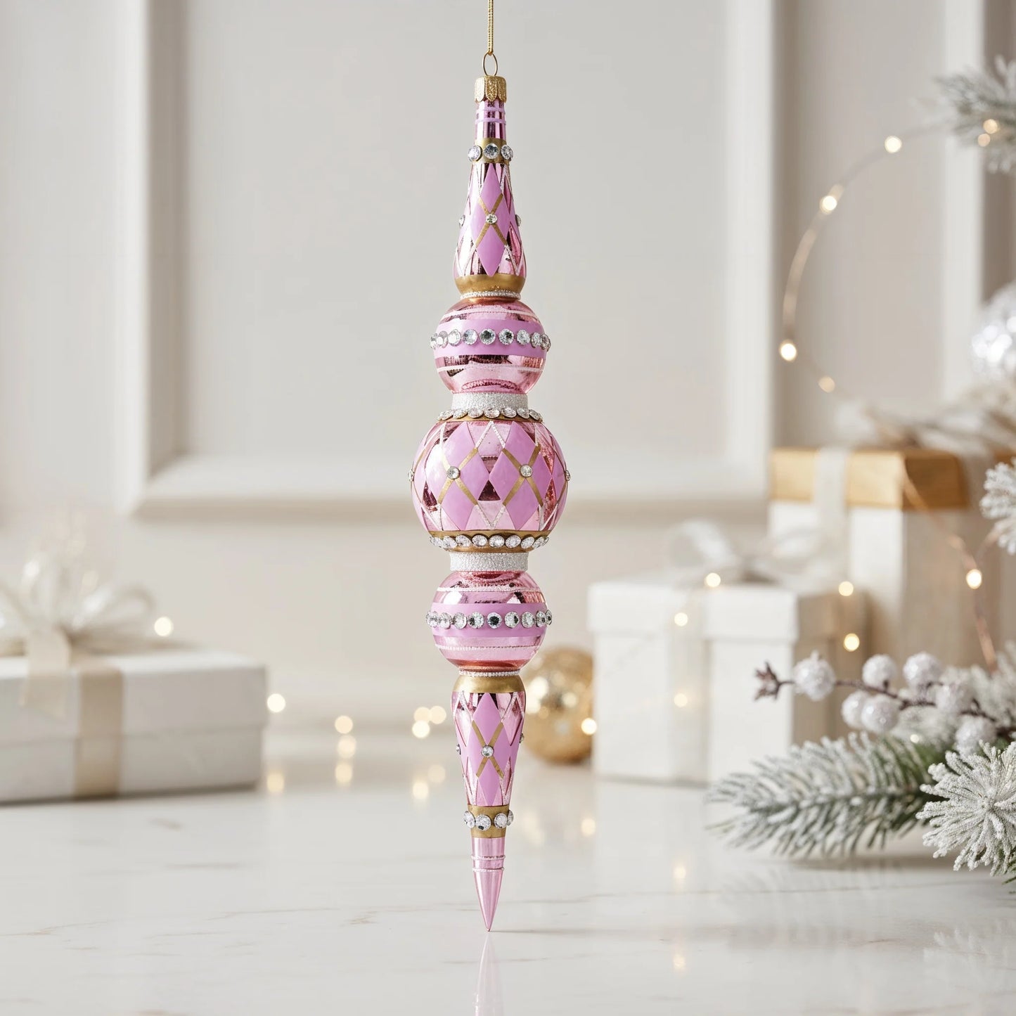Pink harlequin icicle ornament hanging against a soft neutral background with gift boxes and a frosted pine branch, showing its tiered shape, gold diamond pattern, and rhinestone bands catching the light.