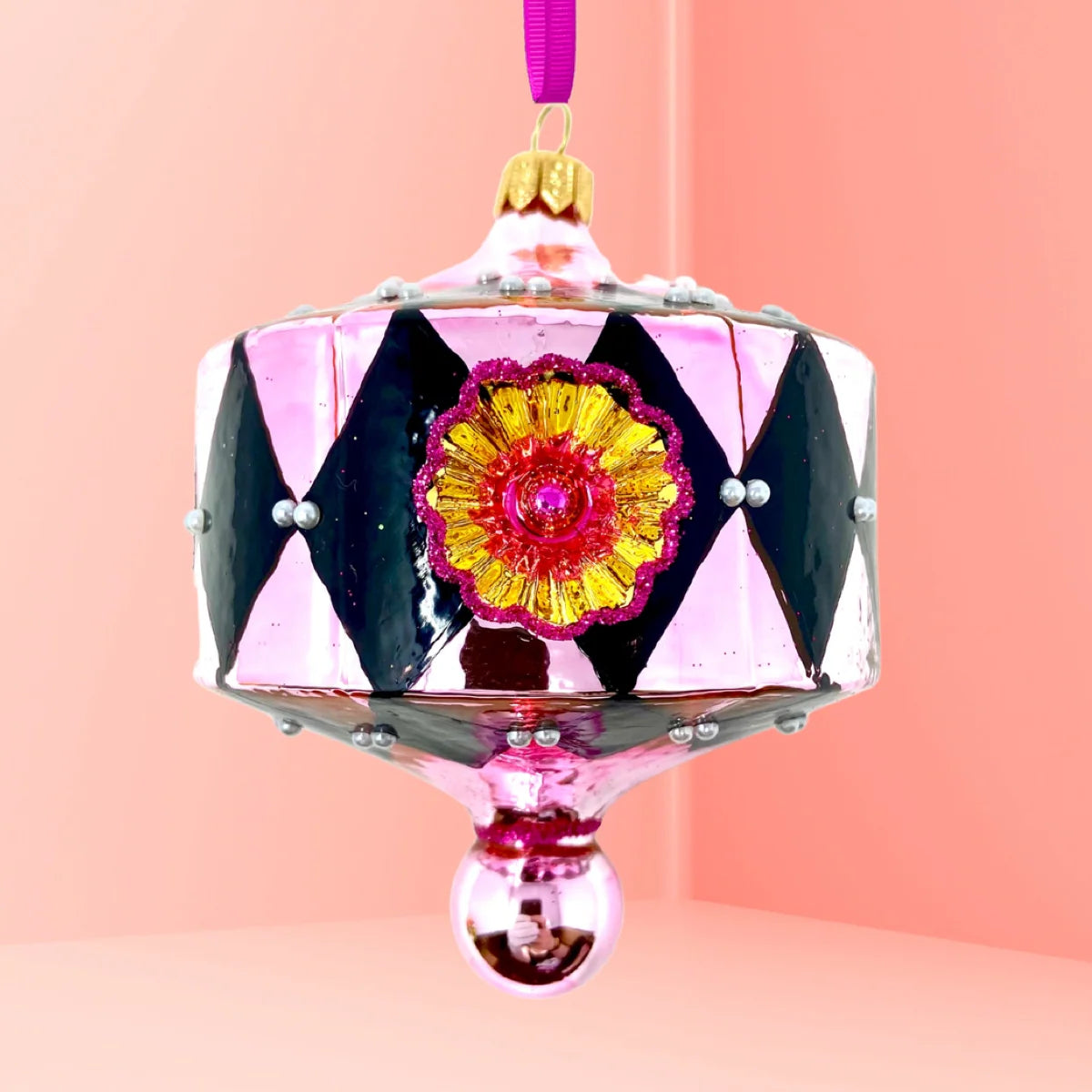Lantern-shaped glass Christmas ornament in blush pink with a black harlequin diamond pattern, faux pearl beads at each diamond point, a gold starburst reflector rimmed in magenta glitter with a red center, a gold cap, and a pink ball drop trimmed in magenta glitter, hung from a purple ribbon.
