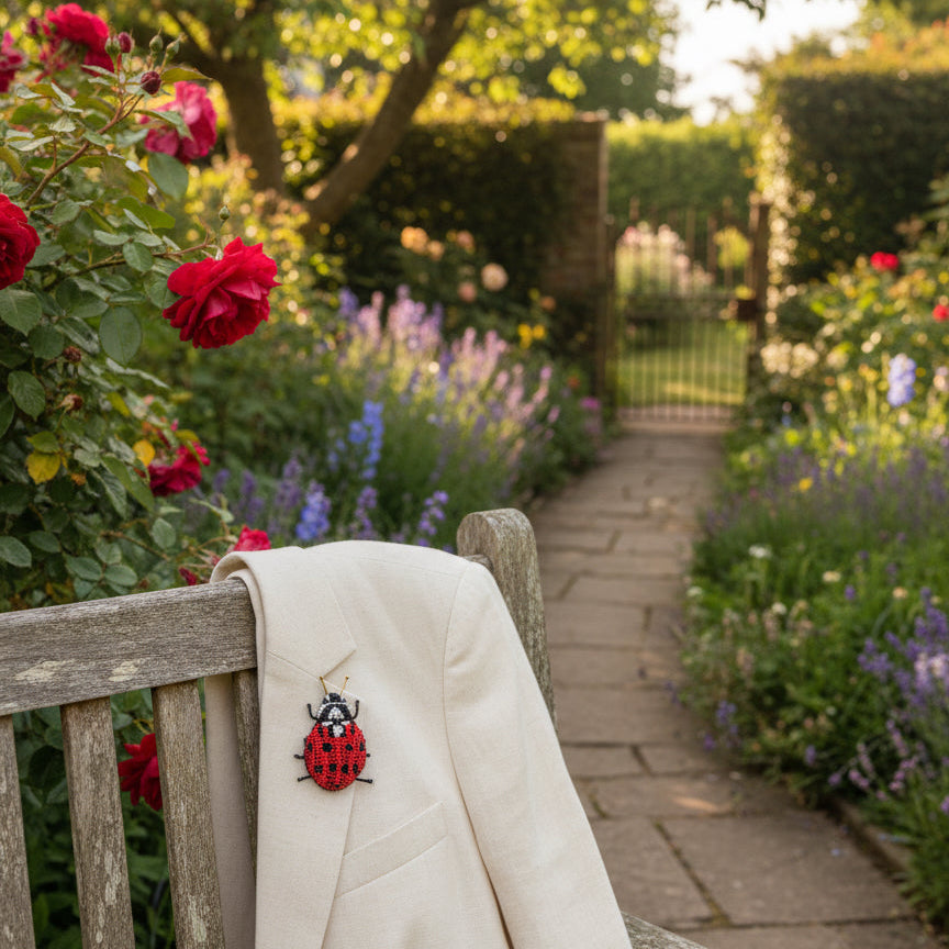 Ladybug brooch pin with a red beaded shell and black spots, pinned to a cream linen jacket draped over a weathered garden bench beside red roses and lavender.
