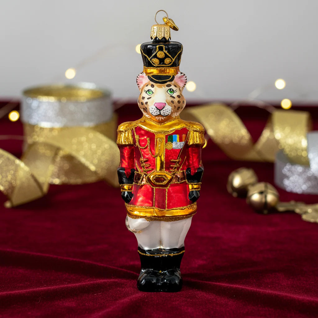 Leopard-shaped glass Christmas ornament dressed as a royal guardsman, wearing a black tall hat with gold trim, a red and gold military jacket with badges and epaulettes, white pants, and black boots, styled among gold ribbon and gift boxes on a maroon velvet surface, hung from a gold ribbon.displays.