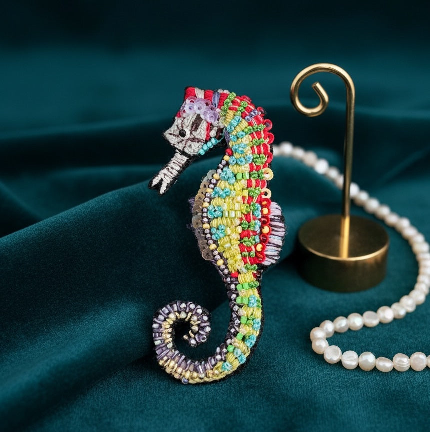 Hand-embroidered seahorse brooch pin with a cream beaded head and snout, a red crest, a body banded in red, chartreuse, yellow, and turquoise beads, a violet striped dorsal fin, and a coiled tail in lavender and gold.