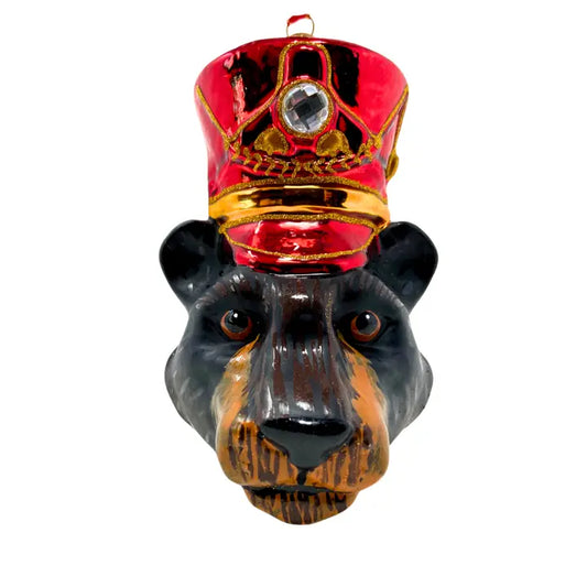 Bear head Christmas ornament in a red ceremonial hat with a gold band, gold trim, and a clear faceted gem, black fur with rust-brown muzzle shading, amber eyes, and a red ribbon loop at the top, shown against a white background.