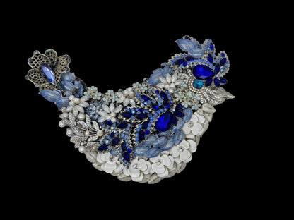 Jeweled blue bird sculpture in side profile, adorned with sapphire and sky-blue rhinestones, pearl accents, enameled flowers, and silver beak, displayed on an easel. Reverse side is painted black. Handmade one of a kind.
