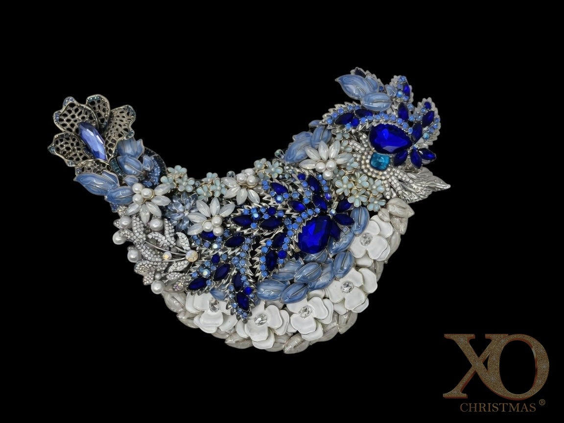 Jeweled blue bird sculpture in side profile, adorned with sapphire and sky-blue rhinestones, pearl accents, enameled flowers, and silver beak, displayed on an easel. Reverse side is painted black. Handmade one of a kind.