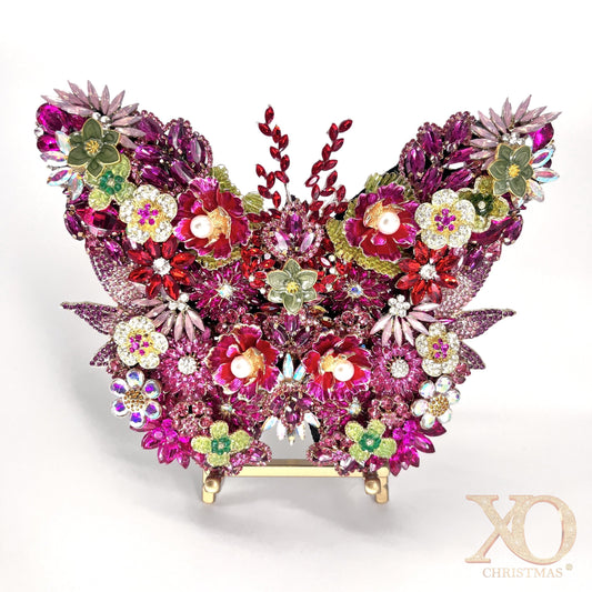 Pink Jeweled Butterfly Sculpture densely layered with fuchsia and magenta rhinestones, pearl-center floral brooches, aurora borealis accents, and sage green enamel flowers, displayed on a gold easel stand against a white background.