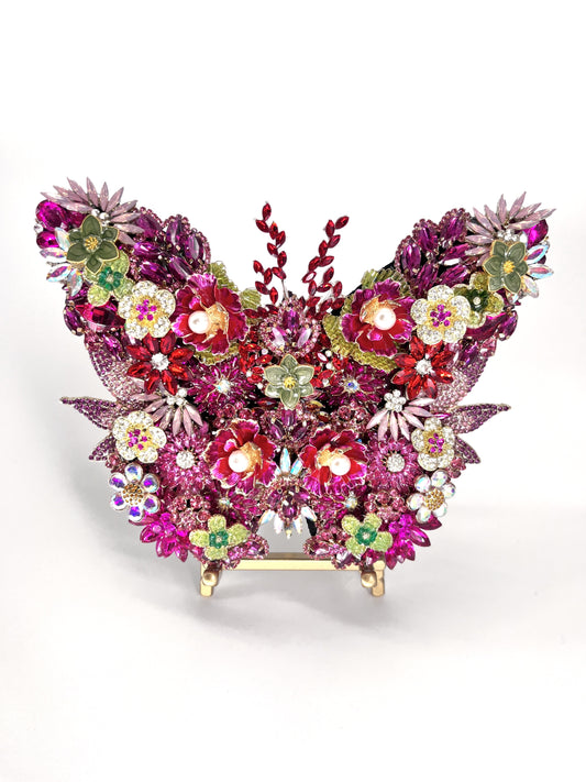 Pink Jeweled Butterfly Sculpture densely layered with fuchsia and magenta rhinestones, pearl-center floral brooches, aurora borealis accents, and sage green enamel flowers, displayed on a gold easel stand against a white background.