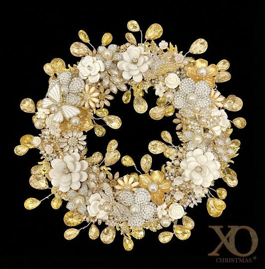 A circular jeweled wreath on a solid black background, covered with white enamel flowers, faux pearl flowers, gold metallic flowers, an enamel butterfly, jeweled hummingbird, dragonfly and miniature cream roses, with faceted champagne teardrop rhinestones hanging from thin wires around both the outer edge and the inner circle.
