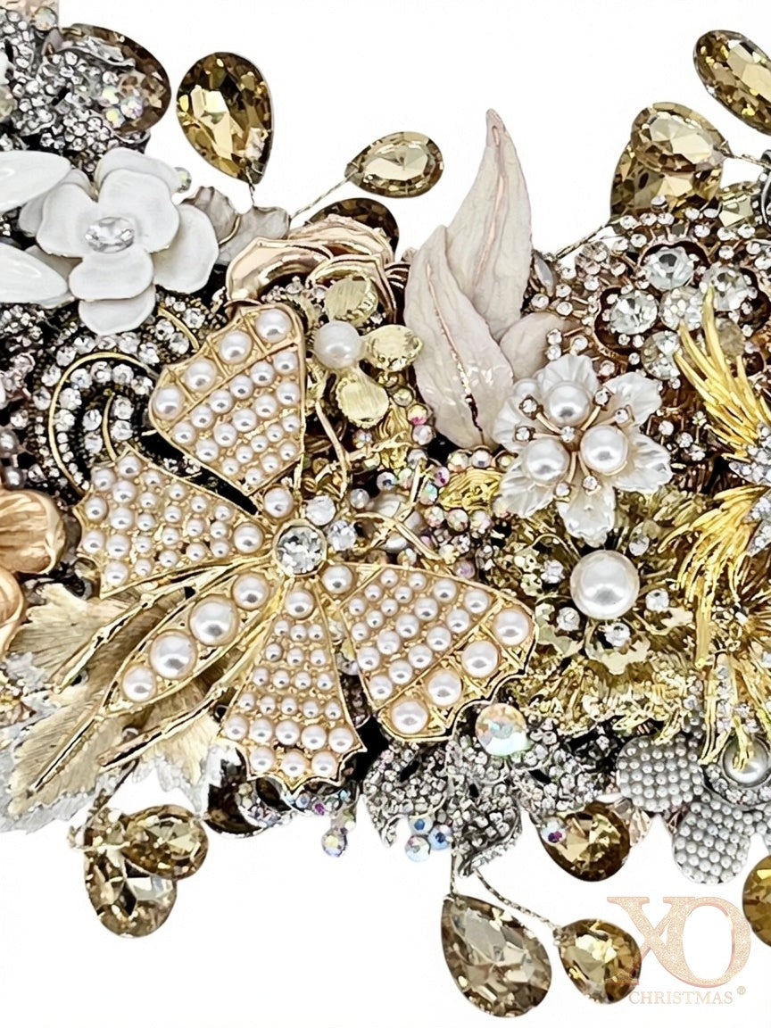 A close-up detail of a jeweled wreath showcasing a golden butterfly in faux pearl-studded accents, white enamel florals, gold filigree leaves, champagne and aurora borealis accent