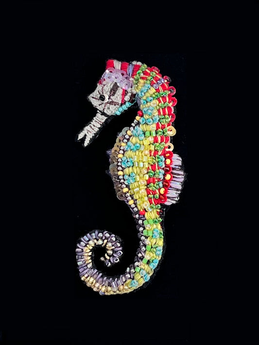Hand-embroidered seahorse brooch pin with a cream beaded head and snout, a red crest, a body banded in red, chartreuse, yellow, and turquoise beads, a violet striped dorsal fin, and a coiled tail in lavender and gold.