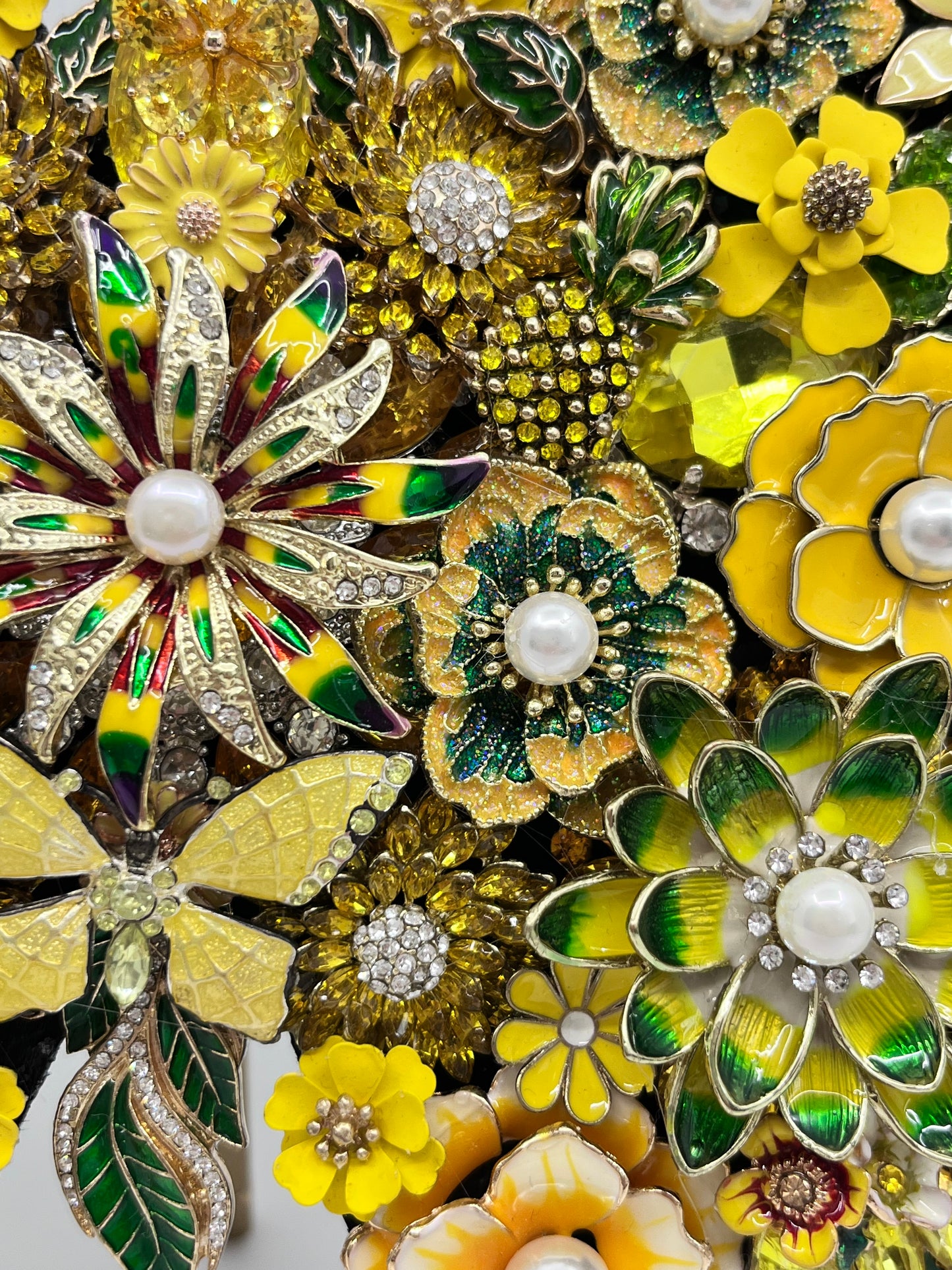 A close-up of multiple vintage-style flower brooch pins in yellow, green, white, and gold tones, arranged tightly together. The brooches feature enamel petals, rhinestone accents, and pearl-like centers. Handmade in the USA.