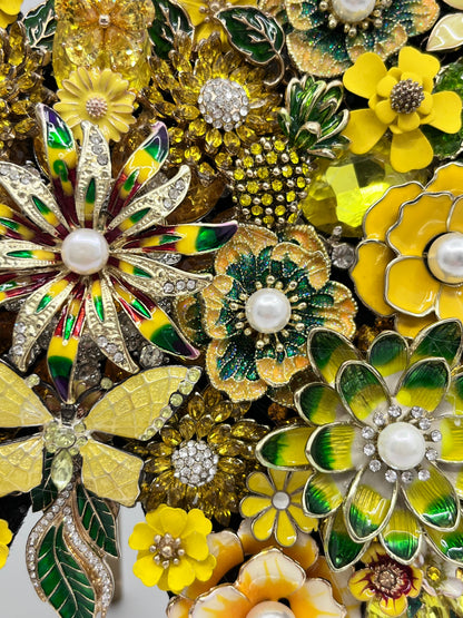 A close-up of multiple vintage-style flower brooch pins in yellow, green, white, and gold tones, arranged tightly together. The brooches feature enamel petals, rhinestone accents, and pearl-like centers. Handmade in the USA.