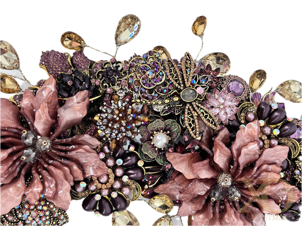 Close-up of an ornate jeweled wreath with two large dusty-rose enamel flowers, a central gold-filigree butterfly with rose-gold and rhinestone body, and clusters of purple and pink rhinestones, faux pearls, and dangling champagne teardrops.