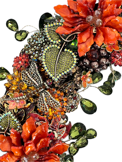 A close-up jeweled floral and leaf embellishments featuring orange and red flowers with champaign breads in center with gold accent, green teardrop rhinestones accenting the outside and inside of wreath, jeweled rhinestone dragonfly 