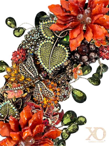 A close-up jeweled floral and leaf embellishments featuring orange and red flowers with champaign breads in center with gold accent, green teardrop rhinestones accenting the outside and inside of wreath, jeweled rhinestone dragonfly 