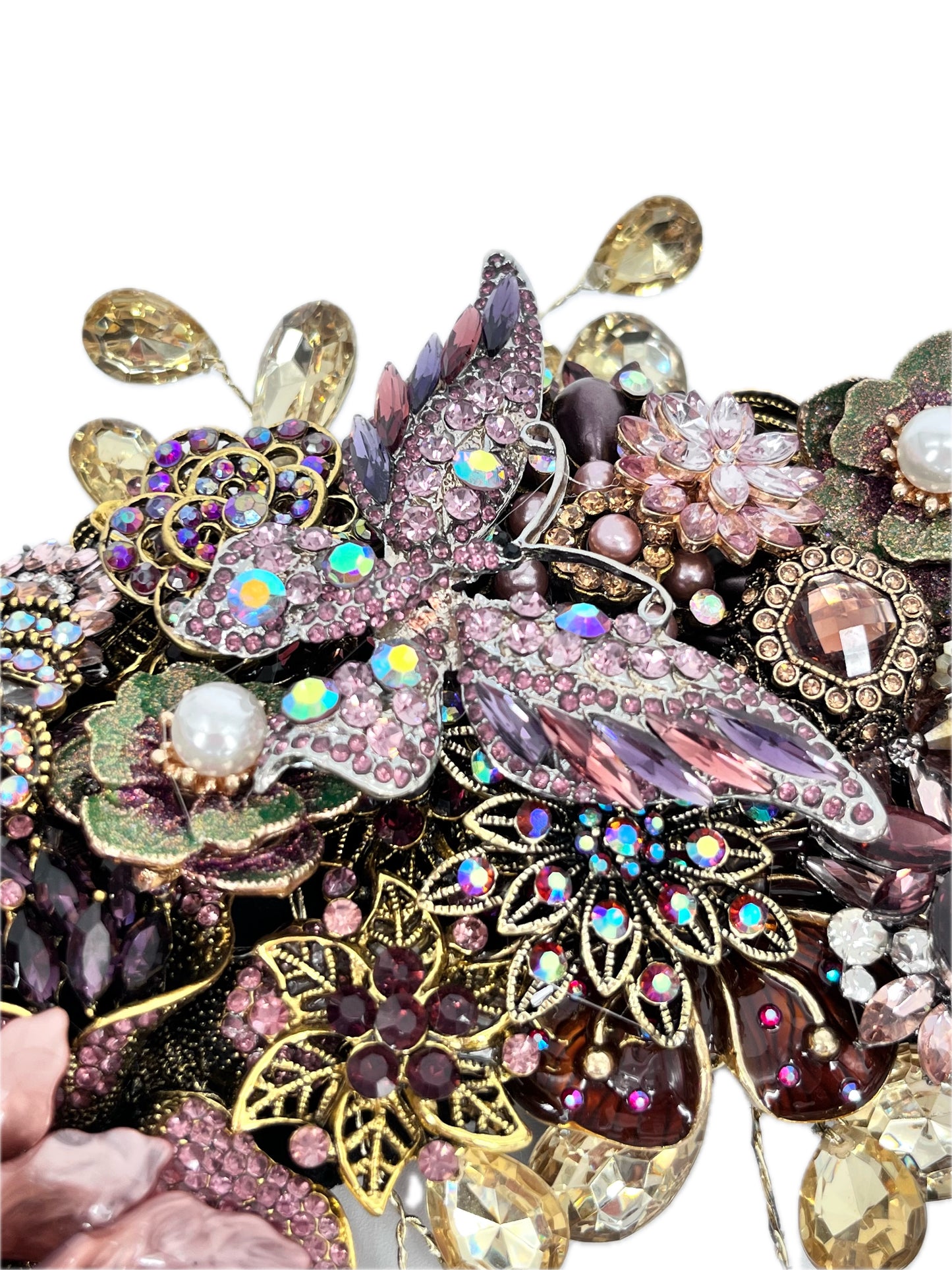 Close-up of an ornate jeweled wreath section centered on a large pink-and-purple rhinestone butterfly with iridescent accents, surrounded by floral motifs, faux pearls, green enamel leaves, purple and amber gems, gold filigree, and dangling champaign teardrop rhinestones.