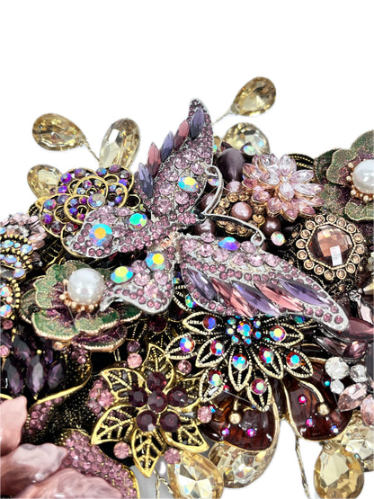 Close-up of an ornate jeweled wreath section centered on a large pink-and-purple rhinestone butterfly with iridescent accents, surrounded by floral motifs, faux pearls, green enamel leaves, purple and amber gems, gold filigree, and dangling champaign teardrop rhinestones.
