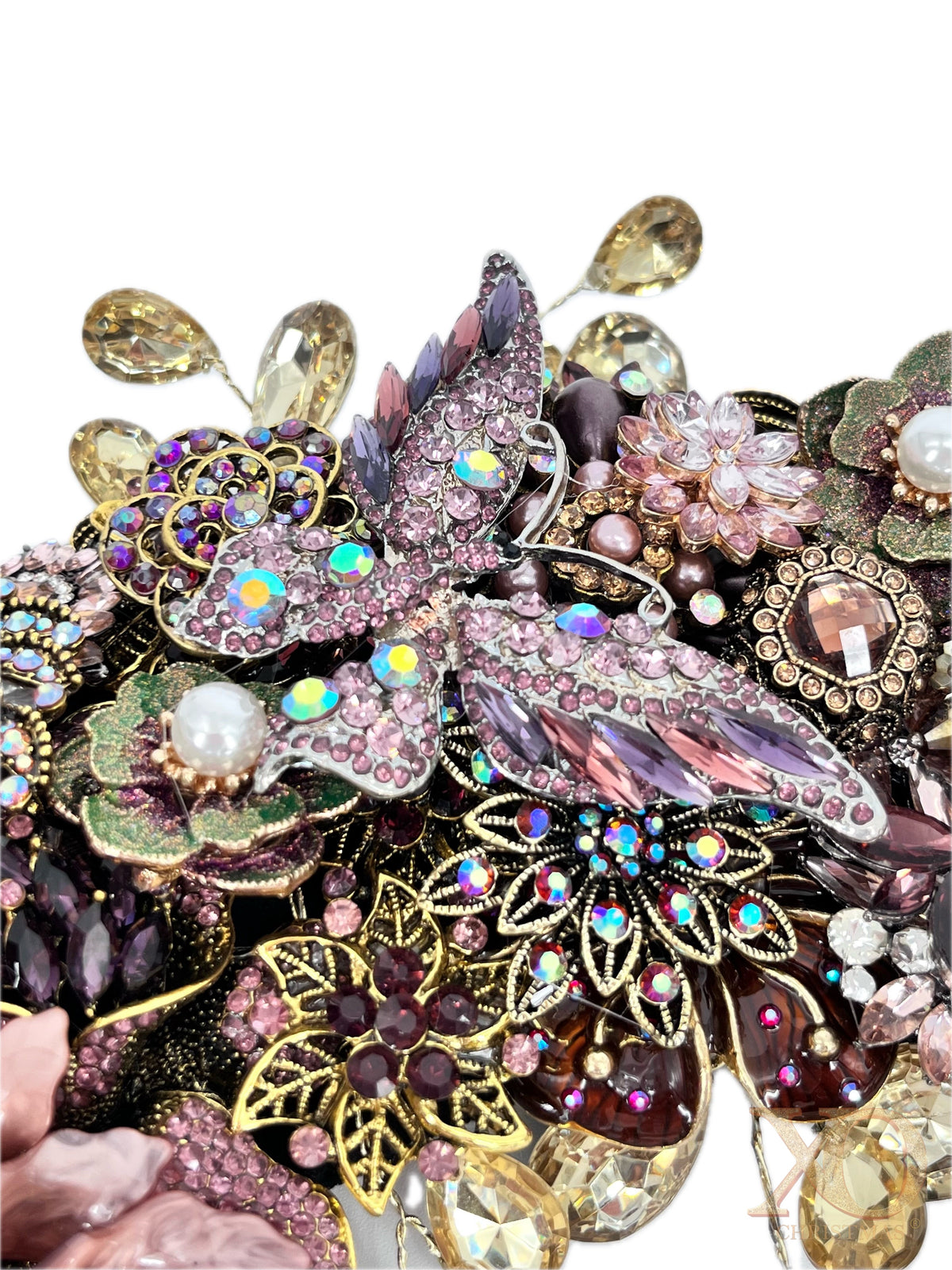 Close-up of an ornate jeweled wreath section centered on a large pink-and-purple rhinestone butterfly with iridescent accents, surrounded by floral motifs, faux pearls, green enamel leaves, purple and amber gems, gold filigree, and dangling champaign teardrop rhinestones.