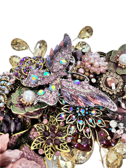 Close-up of an ornate jeweled wreath section centered on a large pink-and-purple rhinestone butterfly with iridescent accents, surrounded by floral motifs, faux pearls, green enamel leaves, purple and amber gems, gold filigree, and dangling champaign teardrop rhinestones.
