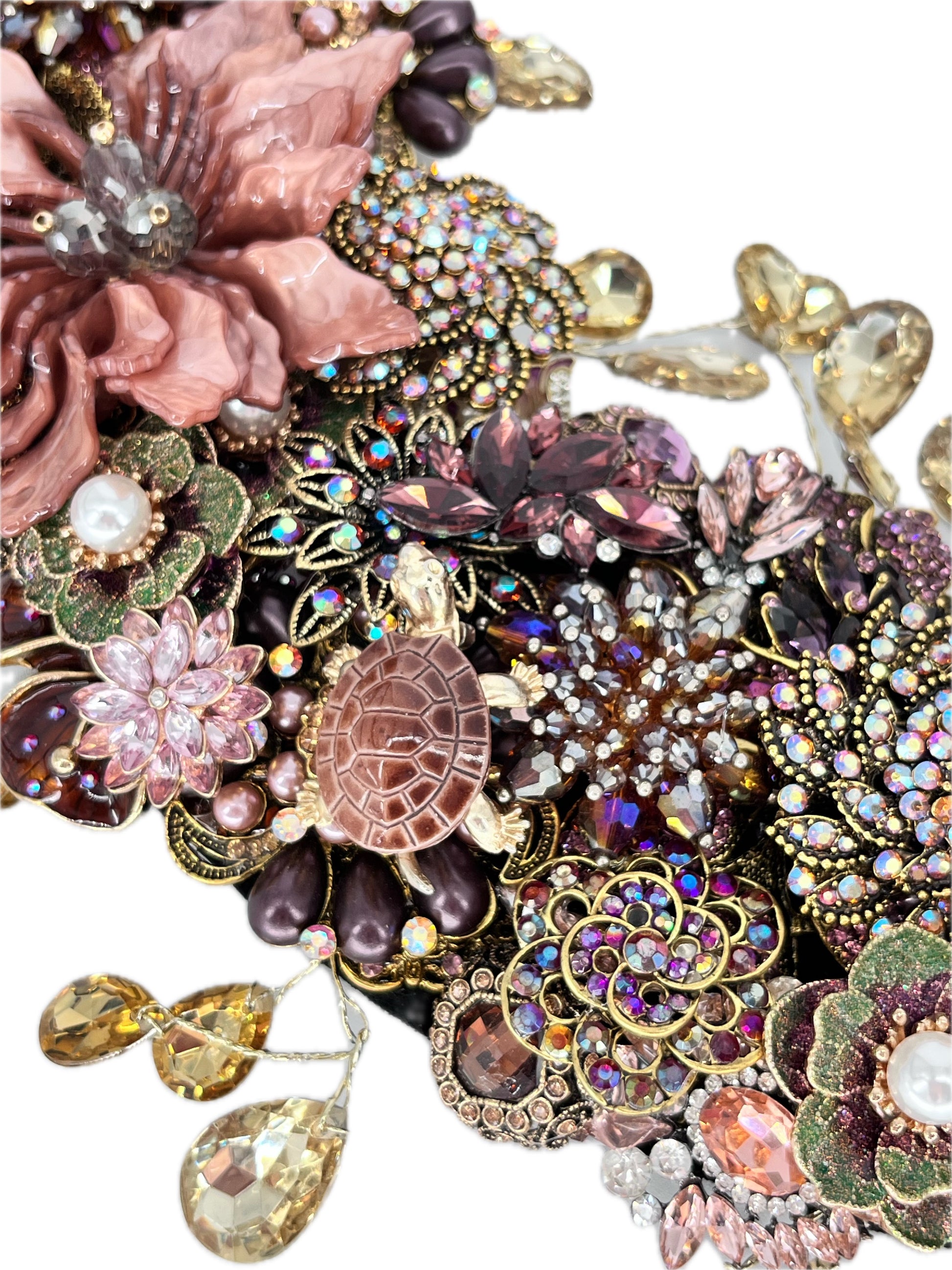 Close-up of an ornate jeweled floral wreath section with a large dusty-pink layered flower, a small brown turtle with a segmented shell, clusters of purple, pink, and iridescent rhinestones, faux pearl accents, gold filigree, and dangling champagne teardrop rhinestones.