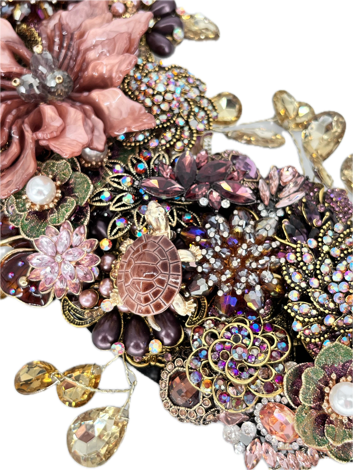 Close-up of an ornate jeweled floral wreath section with a large dusty-pink layered flower, a small brown turtle with a segmented shell, clusters of purple, pink, and iridescent rhinestones, faux pearl accents, gold filigree, and dangling champagne teardrop rhinestones.