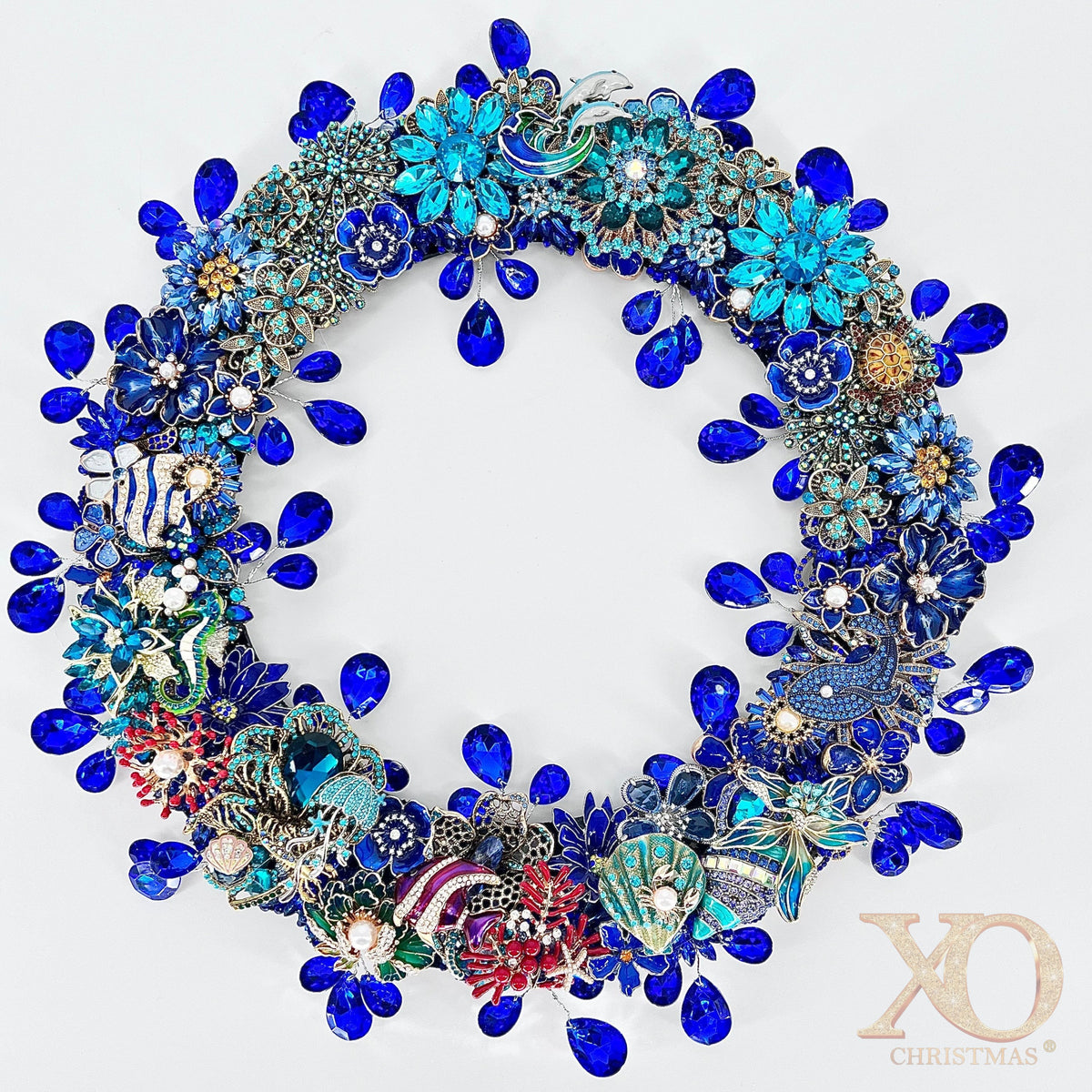 Round jeweled brooch wreath densely covered in cobalt-blue faceted teardrop rhinestones and turquoise enamel flowers set in silver and gold-tone metal. Sea-themed accents include a white and light-blue dolphin at the top, a blue-green seahorse, scallop shells, red coral, and scattered white pearls, on a white background.