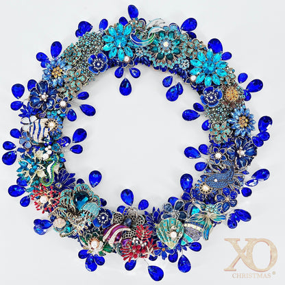 Round jeweled brooch wreath densely covered in cobalt-blue faceted teardrop rhinestones and turquoise enamel flowers set in silver and gold-tone metal. Sea-themed accents include a white and light-blue dolphin at the top, a blue-green seahorse, scallop shells, red coral, and scattered white pearls, on a white background.
