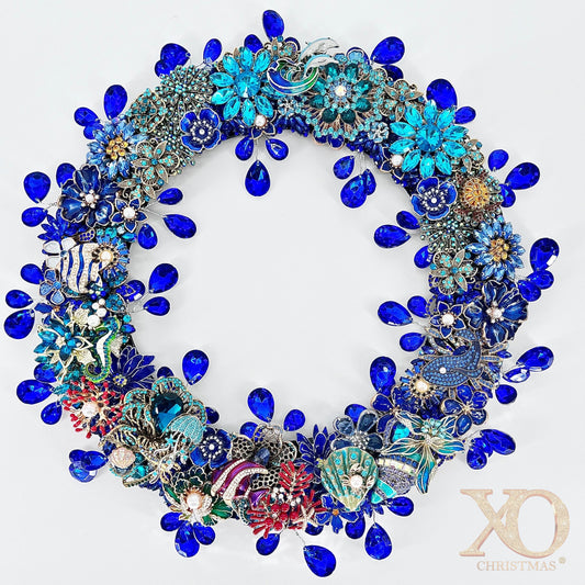 Round jeweled brooch wreath densely covered in cobalt-blue faceted teardrop rhinestones and turquoise enamel flowers set in silver and gold-tone metal. Sea-themed accents include a white and light-blue dolphin at the top, a blue-green seahorse, scallop shells, red coral, and scattered white pearls, on a white background.