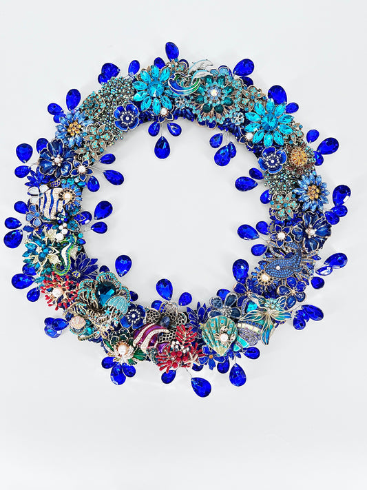 A circular, highly embellished jeweled ocean wreath featuring vibrant blue teardrop rhinestones, turquoise floral clusters, and various colorful vintage-style sea-themed and marine life brooches, set against a plain white background.
