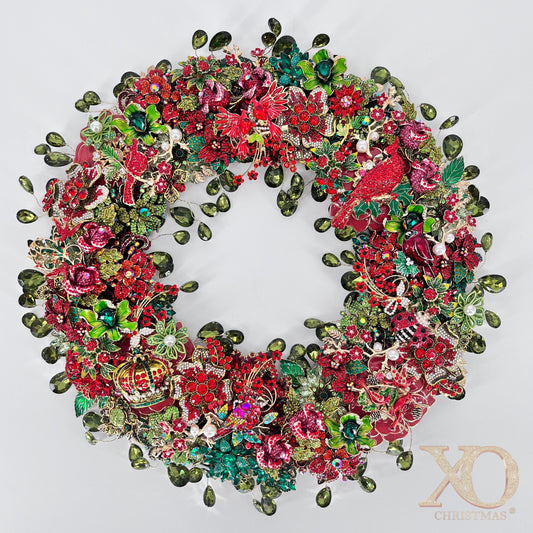 Circular holiday wreath densely covered in costume jewelry, featuring several red cardinal birds, sparkling red and pink rhinestone flowers, green enamel leaves, white pearls, gold-tone metal accents, and dangling olive-green teardrop crystals against a plain white background.