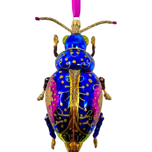 Beetle-shaped glass Christmas ornament in cobalt blue with magenta and black shading, gold glitter scrollwork and dot detailing, gold antennae and legs, hung from a pink ribbon.