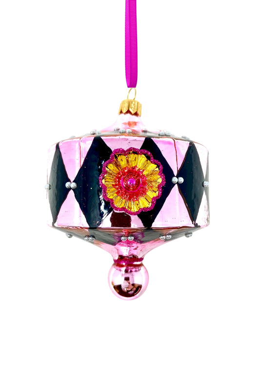 On a white background Lantern-shaped glass Christmas ornament in blush pink with a black harlequin diamond pattern, faux pearl beads at each diamond point, a gold starburst reflector rimmed in magenta glitter with a red center, a gold cap, and a pink ball drop trimmed in magenta glitter, hung from a purple ribbon.