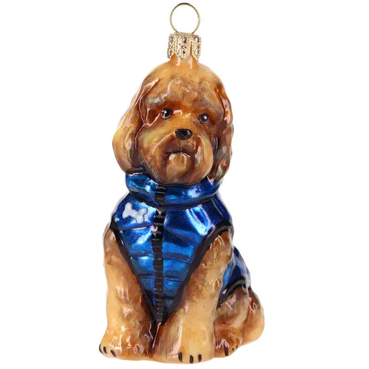 Glass Labradoodle Christmas ornament with tan curly fur, wearing a shiny blue puffer vest with a zipper and white bone patch, hanging on a lit Christmas tree.