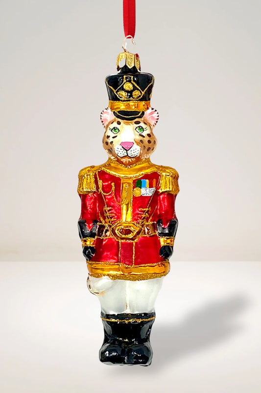 Leopard-shaped glass Christmas ornament dressed as a royal guardsman, wearing a black tall hat with gold trim, a red and gold military jacket with badges and epaulettes, white pants, and black boots, against a neutral background, hung from a red ribbon.