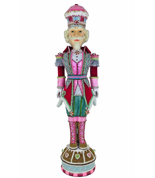 Nutcracker soldier figure with a pink frosted cupcake-shaped hat topped with a peppermint swirl, white curled hair, and a long pointed white beard, wearing a red velvet coat over a pink satin vest and light blue diamond-quilted panel, with silver tinsel epaulettes, peppermint candy appliqués, teal trousers, and pink striped boots, standing on a round gingerbread-style base decorated with icing and candy hearts.