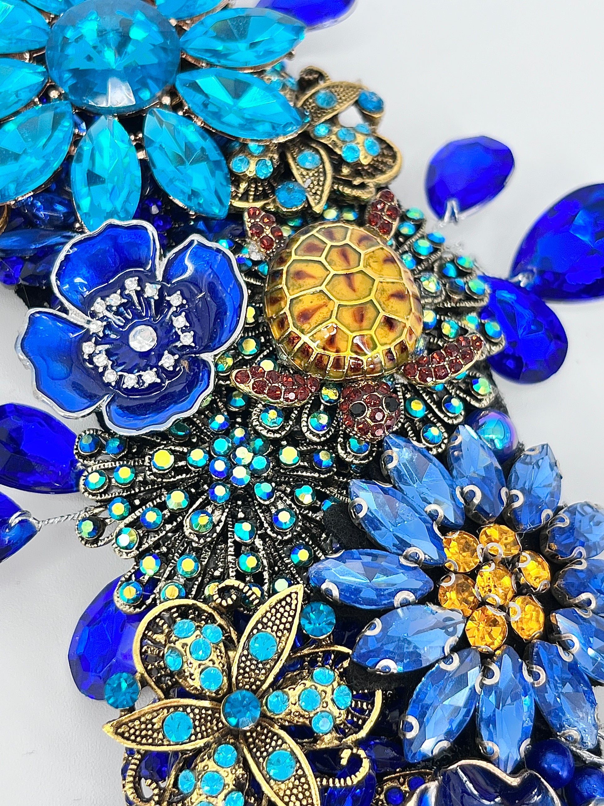 Close-up detail of a jeweled ocean wreath featuring a sea turtle, blue and teal rhinestone flowers, sapphire rhinestone marquise radiating from the outer edge and inner circle, and gold accents.