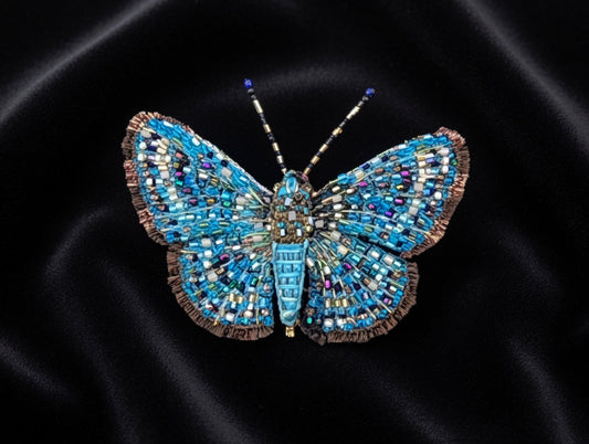 Hand-embroidered butterfly brooch pin with wings spread in a mosaic of turquoise, cobalt, and navy bugle beads flecked with white, gold, and magenta, edged in a bronze fringed border, with a ridged turquoise body and gold and black banded antennae tipped in blue.