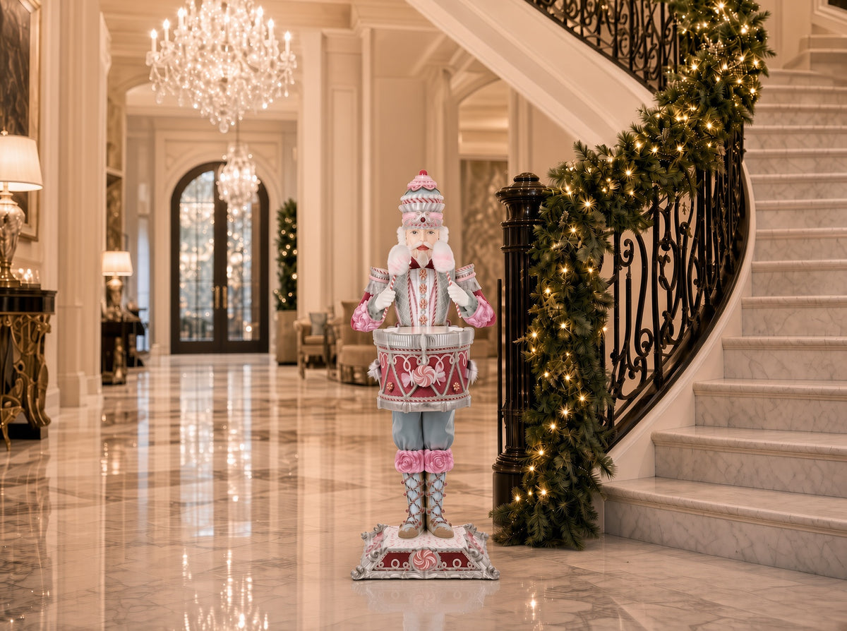 Katherine's Collection Commander Cupcake Nutcracker Server displayed in a marble entryway beside a garland-wrapped staircase, showing the layered cake hat, cotton candy-style drumsticks, and red peppermint-patterned drum in a styled holiday setting.