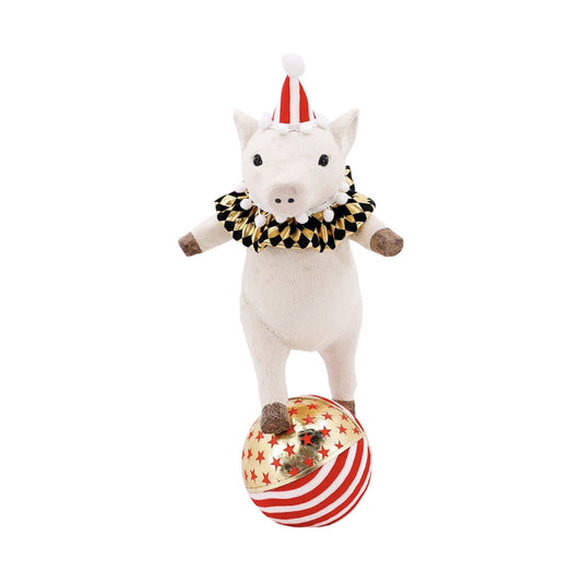 Cream-colored pig decoration with a soft velvety finish standing upright on a ball with arms outstretched, wearing a red-and-white striped party hat with white pom-poms and a black-and-gold harlequin ruffled collar; the ball is metallic gold with red stars on top and red-and-white stripes below.