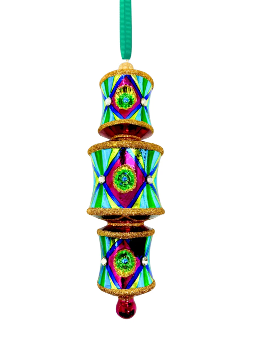 Three-tiered barrel-shaped glass ornament with red and gold glitter banding at the top and bottom of each tier, fuchsia front panels centered on blue reflectors casting a green glow onto yellow, faceted crystal accents, alternating yellow-green and blue striped side panels within a dark blue border, red connecting rings between tiers, and a fuchsia teardrop drop.
