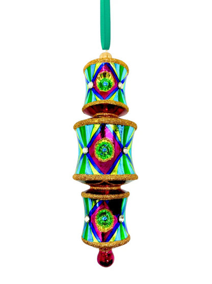 Three-tiered barrel-shaped glass ornament with red and gold glitter banding at the top and bottom of each tier, fuchsia front panels centered on blue reflectors casting a green glow onto yellow, faceted crystal accents, alternating yellow-green and blue striped side panels within a dark blue border, red connecting rings between tiers, and a fuchsia teardrop drop.