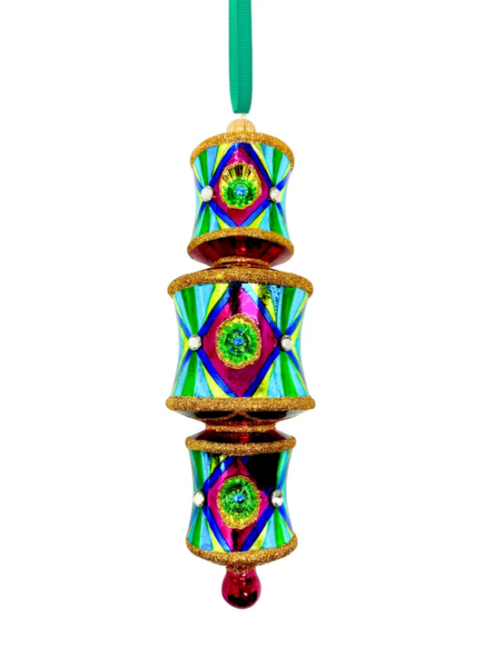 Three-tiered barrel-shaped glass ornament with red and gold glitter banding at the top and bottom of each tier, fuchsia front panels centered on blue reflectors casting a green glow onto yellow, faceted crystal accents, alternating yellow-green and blue striped side panels within a dark blue border, red connecting rings between tiers, and a fuchsia teardrop drop.