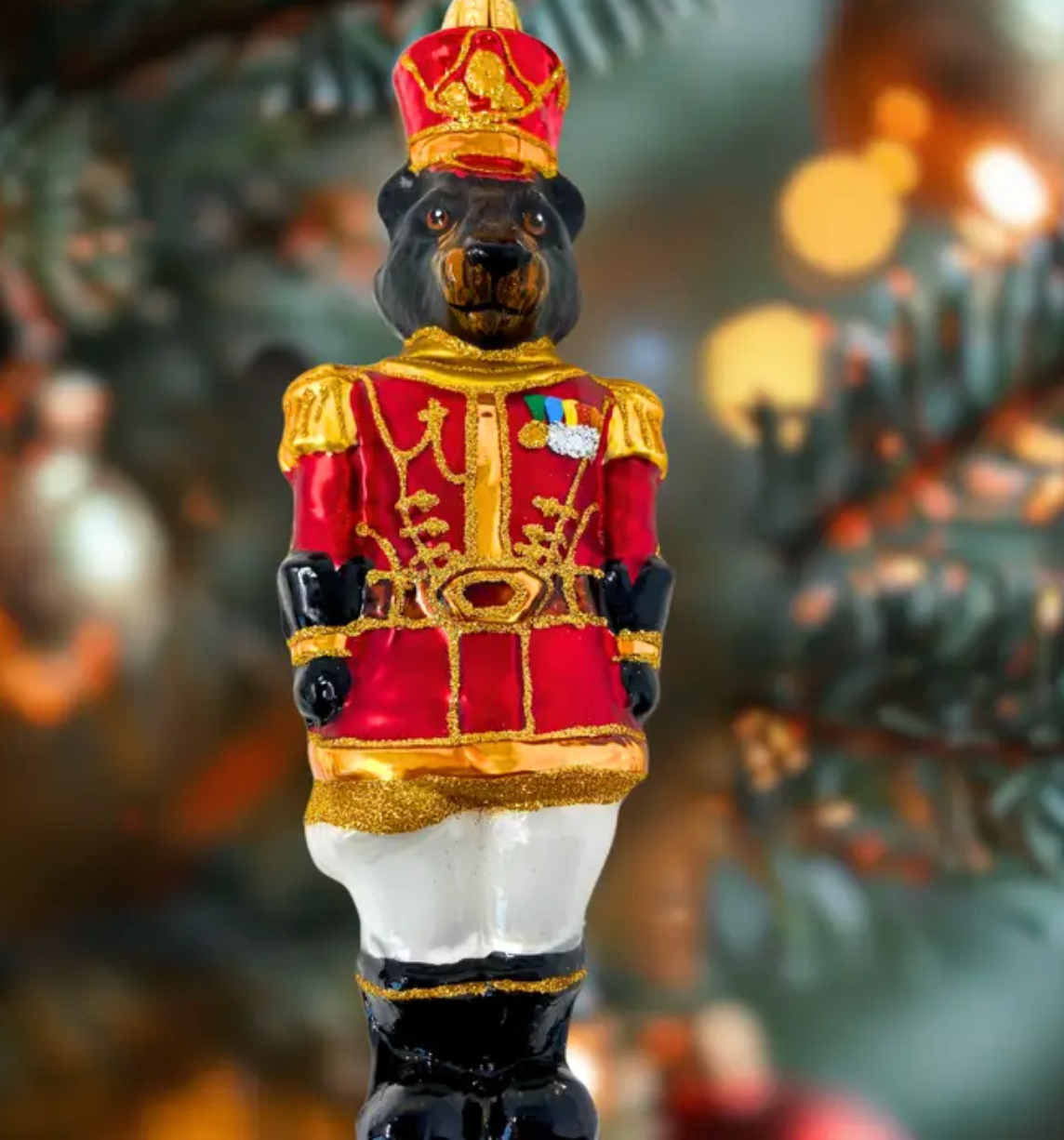 Bear-headed nutcracker soldier ornament in a red and gold ceremonial military coat with gold epaulettes and a striped ribbon medal, tall red guard's hat with a gold band and crest, white trousers, and black boots, shown against a softly blurred, light-filled background.