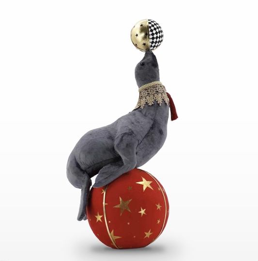 Gray seal figure with a velvety texture perched on a red ball decorated with gold stars and a gold stripe. It balances a smaller ball on its nose that is half gold with dark stars and half black-and-white checkered, and it wears a scalloped gold lace collar with a red tassel.