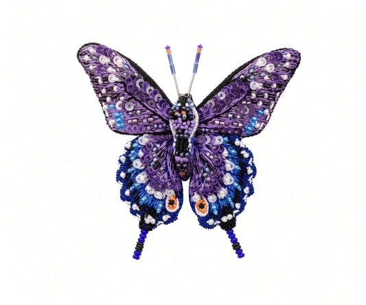 Close view of a hand-embroidered swallowtail butterfly brooch pin showing overlapping purple sequin upper wings, deep blue lower wings dotted in white beads, two orange eyespots, black wing edging, and fine beaded tails and antennae on a white background